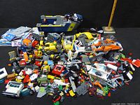 Wide view of mixed LEGO bricks, partial builds, and some vehicle parts laid out in a pile including yellow and blue pieces, a boat-like structure, wheels, and various small components.