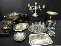 Full lot showing all silver-plated dinnerware pieces, glass condiment holders, goblets, and the five-arm silver-plated candelabra against a black backdrop.