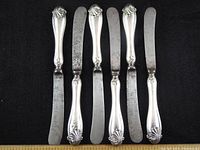Set of 6 butter knives with sterling silver handles and blades showing tarnish, arranged on black background with ruler for scale.
