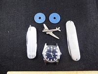 Photo showing two blue WWII Canadian meat ration tokens, vintage Timex watch case, two silver-tone pocket multi-tools, and a small silver airplane pin arranged on dark fabric