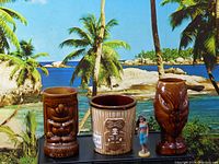 Three vintage tiki mugs and a small figurine against a tropical island background