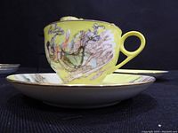 Close-up image showing a porcelain tea cup with a yellow background and intricate traditional Chinese figure design on the outer surface, along with the matching saucer.