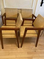 Four compact Mid Century Modern style armchairs grouped close, showing teak frames and buttercream vinyl cushions.