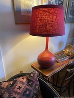 Red ceramic lamp turned on with textured red linen shade emitting warm light on wooden console table next to patterned pillow.