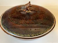 Close-up photo of the pottery lidded dish showcasing the dark brown glaze with cinnamon and moss green decorative details, and the twisted rope handle on the lid.