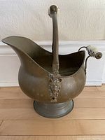 Front view of large brass pot showing spout, central handle, and side ceramic handle with decorative blue and white pattern.