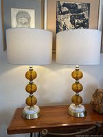 Pair of lit lamps on wooden table with white drum shades, artwork in background