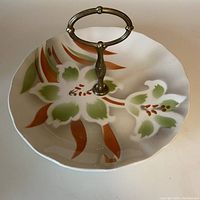 Side angle showing cinnamon and olive green floral design, scalloped ceramic plate with tall removable brass handle.