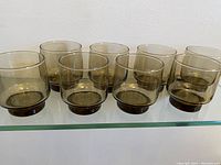 Eight smoky brown lowball glasses arranged in a row on a glass shelf.