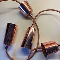 Two solid copper cylindrical hanging light shades with attached cords and copper stays.