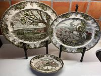 Set of three Johnson Bros. Friendly Village oval serving plates with scenic village designs displayed on stands