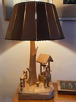 Full view of carved lamp showing wooden slatted lampshade, tree trunk base, man, well, and bucket on a carved platform.