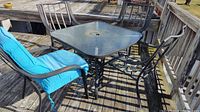 Full view of the 5 piece deck furniture set on a wooden deck showing four metal chairs with blue cushions and a square glass top table with black metal frame.