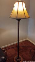 Full view of lit bronze finish floor lamp with cream fabric pleated shade with brown trim on edges.