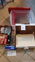 Photo showing clear plastic file bin with red handles, Cannon P26-DH calculator, box of envelopes, stapler, and small items spread on floor.