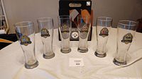 Six tall clear glass commemorative mugs displaying Pittsburgh Steelers Super Bowl logos for the years 1975, 1976, 1979, 1980, 2006, 2009 arranged on a white cloth.