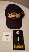 Black 2009 City of Champions Pittsburgh hat with embroidered lettering placed above the black porcelain Pittsburgh Steelers light switch cover showing logo and text.