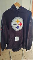 Black hooded pullover sweatshirt with large Steelers logo on the chest, hanging on a hanger.