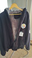 Black front zip Steelers sweatshirt shown hung on wooden hanger. Open zipper reveals soft grey inner lining and hood interior.