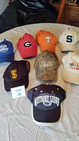 Photo showing nine sports hats arranged on a white table with various college and brand logos visible.