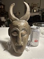 Front view of a hand-carved wooden tribal mask with two large curved horns, next to a soda can for size comparison.