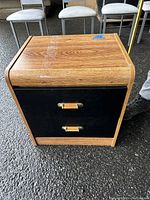 Front and angled view of the wooden two drawer bedside table showing the wood grain top and black drawer fronts with brass handles.