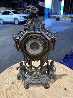 Front view of brass ornate clock with four small human figurines supporting the frame and detailed finial on top.