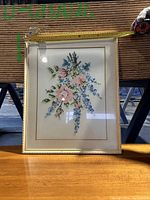 Cross-stitch floral artwork inside a thin gold frame with white mat, laying flat showing approximate width measurement.
