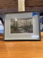 Framed cross-stitch artwork with a landscape view including trees, river, and cabin under cloudy sky, shot under natural light showing reflections on the glass.
