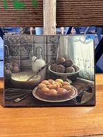 Framed still life print showing a kitchen counter with various food items including eggs, fruit, and utensils in a transparent frame.
