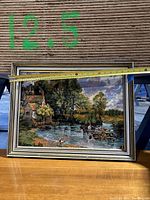 Full view of the framed Petit Point needlework showing a lake with trees, cottage, dock, and people. Measuring tape across the frame shows approximately 22 inches width.