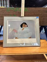 Front view of framed art print titled 'Mele Kalikimaka 1982' featuring a person in white clothing with black hair and pink flower, resting chin on hand, with a gray background and the text 'Mana's Gallery' below.