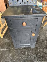 Front-facing image showing black wooden cabinet with drawer and door, wooden knobs, and decorative scalloped back panel with heart-shaped cutouts. Visible wear and surface scratches.