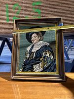 Framed needlepoint stitch artwork depicting a man in Elizabethan costume with ruffled collar and blue-gold attire, measured at about 12.5 inches wide.