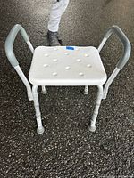 Front view of white plastic shower chair with textured seat, aluminum frame, grey rubber handles and feet on rough floor surface.