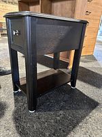 Black wooden side table viewed from an angle showing the drawer, lower shelf, and legs.