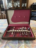 Open presentation box reveals silver flatware including forks, knives, spoons and napkin rings organized on red fabric.