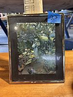 Framed print featuring a woman in garden scenery with flowers, wheelbarrow, and chickens.