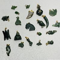 Overhead view of all 20 jade pendants spread on a light patterned background showing various animal and abstract shapes and metal bails for hanging.
