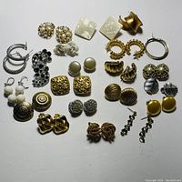 Top-down view of 26 pairs of costume jewelry earrings in various designs, materials, and colors laid out on a white surface.
