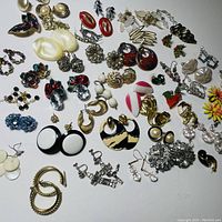 Assortment of costume jewelry earrings laid out on a white surface, showing variety of styles including hoops, dangles, clip-ons, and studs in multiple colors and materials.