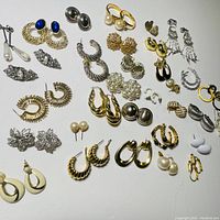 Image of a collection of 28 pairs of costume jewelry earrings displayed on a white surface, showing a variety of styles, colors, and clip-on and post-back closures.