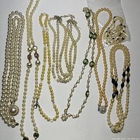 Top-down view of assorted faux pearl costume jewelry pieces arranged flat on a white surface. Visible items include multiple pearl necklaces of various styles, a pair of matching faux pearl earrings, and a small plastic bag containing additional items.