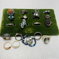 Image showing 16 assorted costume jewelry rings including colored stones, metal bands, and bead bracelet, displayed on and around a green velvet ring holder.