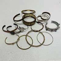 Front view of 14 different cuff bracelets and bangles arranged on a white surface displaying variety in style, texture, and colors