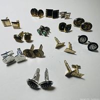 Top view of 12 pairs of men's cufflinks in various designs, shapes and materials laid on white surface.