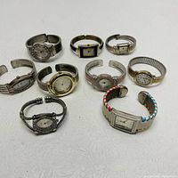 Top-down image of 9 assorted vintage cuff and wrist watches in various metal finishes and face designs on white background