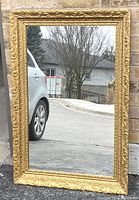 Full view of the large rectangular gilt wall mirror standing outdoors, showing detailed ornate carved frame work and reflective glass surface.