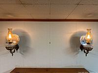 Pair of illuminated brass and glass oil lamp sconces mounted on wall with blown glass reflectors behind the lamps