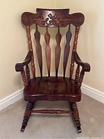Front view of the solid wood rocking chair showing carved floral decorative top, turned spindle backrest and arms, polished wood seat, no cushions.